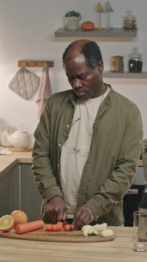 Orta yaşlı Afrikalı bir adamın modern mutfakta yemek masasında durup ahşap tahtada taze havuç keserken blender 'da sağlıklı meyve püresi yaparken orta boy görüntüsü.