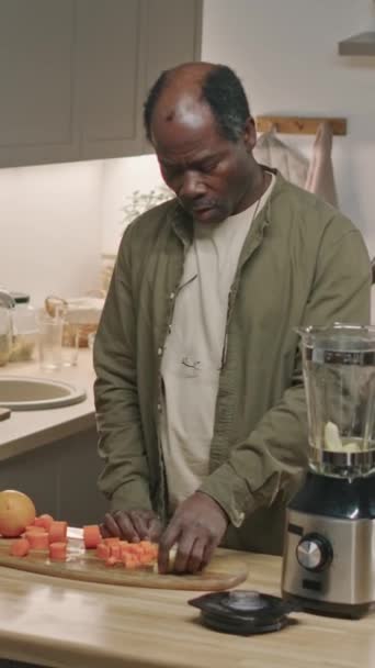 一个退休黑人男子将新鲜胡萝卜和苹果放进搅拌机里 同时在现代厨房做早餐用的健康软糖的垂直中图 — 图库视频影像