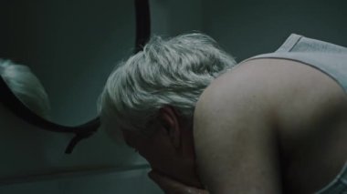 Hüsrana uğramış üzgün, gri saçlı, beyaz bir adam lavaboda yüzünü yıkıyor ve aynadaki yansımasına bakıyor. Soğuk, karanlık banyoda tek başına duruyor.