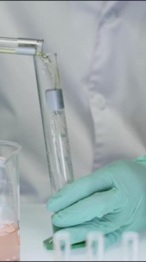 Laboratuvarda kimyasal deney yaparken beyaz laboratuvar önlüğü ve tek kullanımlık eldivenlerle cam test tüpünü yeşil sıvıyla dolduran tanımlanamayan kimyagerin dikey yakın çekimi.