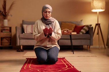 Gündelik giysili genç İslami kadın ve ev ortamında küçük kırmızı halıya diz çökerken avuçlarını açık tutan grye tesettürlü kadın.