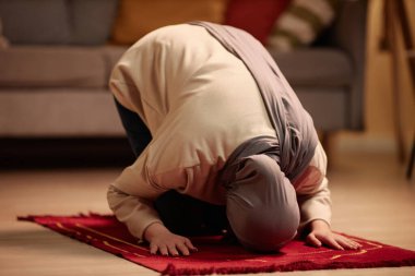Başörtüsü ve bej rengi kazaklı genç Müslüman bir kadın dizlerinin üzerinde durup Allah 'a dua ederken öne eğiliyor.