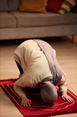 Bej renkli kazaklı ve gri tesettürlü genç Müslüman kadın evde yerde diz çökerek küçük kırmızı halıda dua ediyor ve öne doğru eğiliyor.