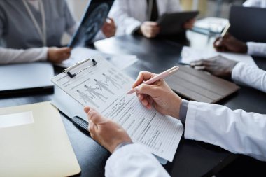 Tıbbi formları grafik ve kişisel bilgilerle dolduran kadın klinisyen bir grup iş arkadaşına karşı masada otururken,
