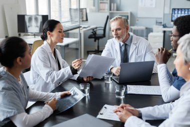 Bir grup kültürler arası meslektaşın önünde masada oturan deneyimli erkek doktora tıbbi belgeler gösteren genç bir klinisyen.