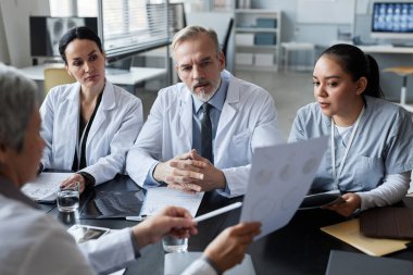 Rapor veya sunum sırasında meslektaşları tarafından tutulan belgelere tıbbi verilere bakan bir grup genç ve olgun klinisyene odaklanın