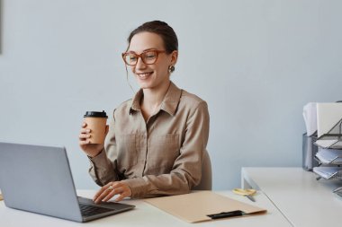 Ofiste dizüstü bilgisayar kullanırken gülümseyen genç bir kadının minimum portresi ve bir fincan kahvenin ve fotokopi alanının keyfi.