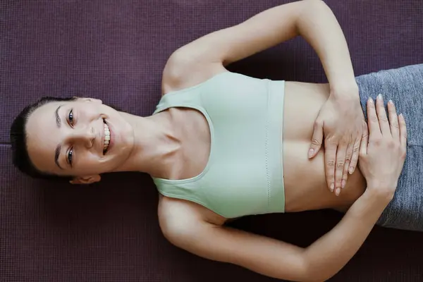 Top view of smiling young woman doing yoga and lying on floor with hands on stomach, copy space