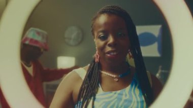 LED yüzüklü genç siyahi kadının parlak y2k kıyafetleri içinde kamera karşısında dans ederken ve çok ırklı bir çift arkadaşının arka planda dumanlı odada dans ederken gülümsediği bir sahne.