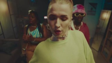 Orta yaşlı, çekici, beyaz, esnek vücutlu sarışın bir kadın. Kamera karşısında baştan çıkarıcı bir şekilde dans ediyor. Arka planda hareket eden birkaç Afrikalı Amerikalı dansçı var.