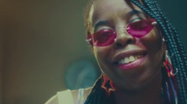 Saçları örgülü, pembe gözlüklü, kameraya poz veren ve el fenerinde dans eden Afro-Amerikan genç bir kadının yavaş yavaş yakın plan portresi.