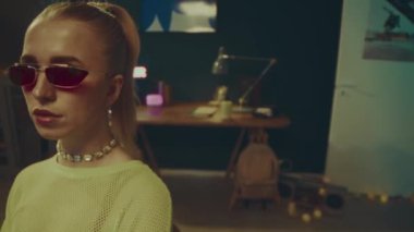 Yüksek açılı, orta ölçekli, çekici, pembe gözlüklü genç bir kadının portresi ve sıcak ışık odasında kameraya poz veren neon sarısı örgü bluzu.