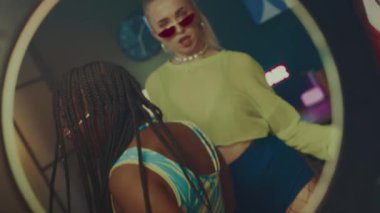 Orta yaşlı Afrikalı Amerikalı genç bir adam ve iki çok ırklı kız. Hepsi parlak Y2K giysiler giyiyorlar. Kameraların önünde dans ediyorlar.