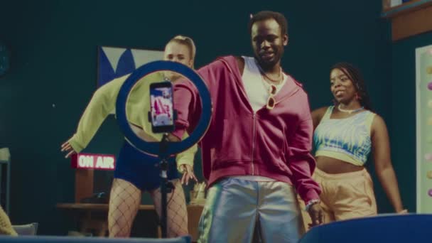 三位20岁出头的多种族朋友穿着Y2K系列服装 在生机勃勃的五颜六色的房间里跳舞 一边用三脚架通过智能手机进行现场直播 他们的速度是中等的 — 图库视频影像