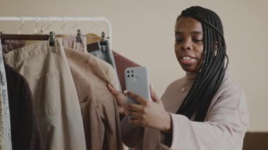 Süslü siyah kadın moda blogcusu askılarda şık giyim eşyalarıyla rafların yanında duruyor ve akıllı telefondan alışveriş vlog 'u kaydediyor.