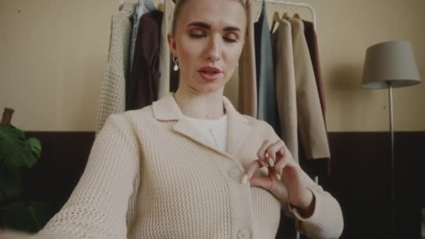 若い白人女性ファッションブロガーのハンドヘルドPovショットは ミニマリストアパートでソーシャルメディアのビデオを録画しながら ボタンで加入者にそれを実証するスタイリッシュなニットカーディガンを身に着けています — ストック動画