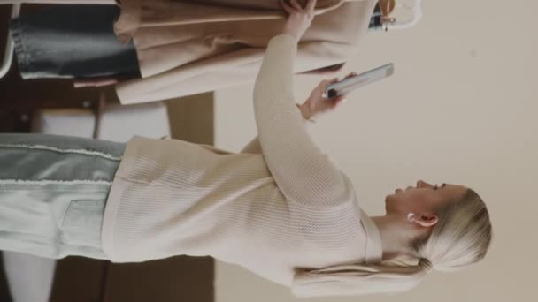 ラックに掛かるトレンディな服のスマートフォンでビデオを撮影する若い白人女性ファッションブロガーの垂直中遅い — ストック動画
