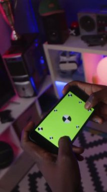 Elinde yeşil krom ekran olan akıllı telefonu tutan ve çevrimiçi oyun oynayan, apartmanın karanlık neon ışıklı odasında duran tanınmayan oyuncunun dikey kırpılmış görüntüsü.