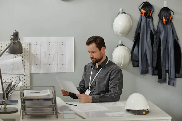 Minimal portrait of bearded male engineer working at desk in office against white wall with workwear in background, copy space