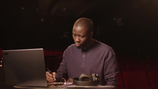 年轻的黑人男喜剧演员坐在黑暗的业余剧场的笔记本电脑旁 一边看着镜头 一边为他的音乐会写笑话 一边拍中肖像画 — 图库视频影像