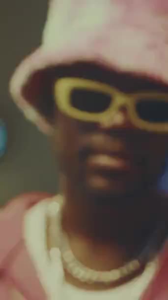 身穿粉色毛绒斗篷帽和黄色眼镜的时髦非洲裔美国嬉皮士男子在室内摆姿势拍照的垂直缩影特写 — 图库视频影像
