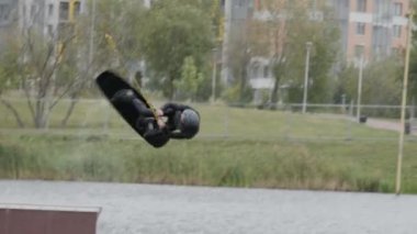 Kafkaslı ve dalgıç kıyafetli beyaz wakeboardcunun sürgülü kayarak suya düşüşünün yavaş çekim görüntüsü.