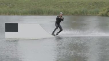 Profesyonel yetişkin beyaz erkek wakeboardcu kaydırağın üzerinden atlarken kablo parkında antrenman yaparken numara yapıyor.