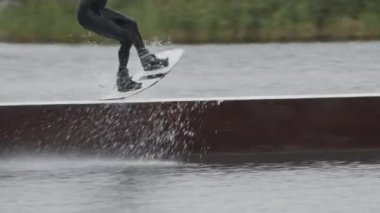 Profesyonel erkek wakeboardcunun kablo parkında sürgülü kayma denemesi yaparken elinde iple dalgıç kıyafeti ve kask tutarken görüntüsü.