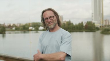 Kameraya poz veren orta yaşlı, beyaz tenli, gözlüklü, elleri su kenarında katlanmış bir adamın orta yavaş portresi.