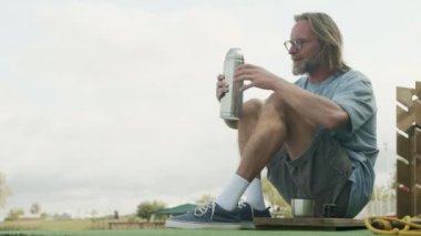 Beyaz yetişkin bir adam suni yeşil çimenlik paspasın üzerinde oturuyor ve termostan sıcak çay içiyor.