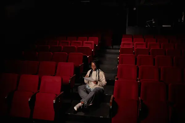 Minimal wide angle view at female African American artist rehearsing lines sitting alone in theater audience with low light copy space