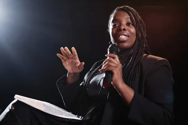 Minimal portrait of African American woman speaking to microphone on stage with dark background copy space