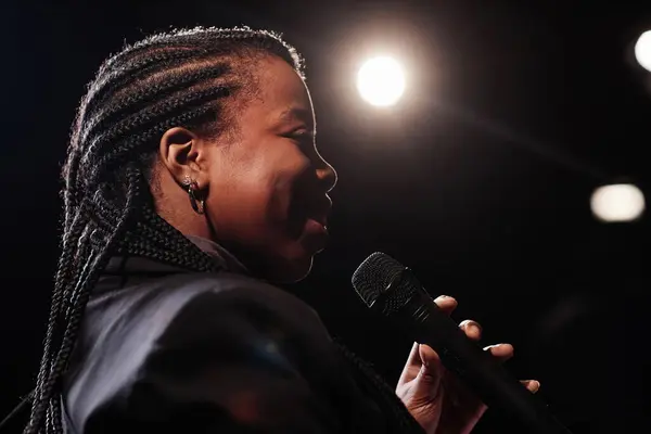 Side view portrait of smiling Black woman holding microphone and performing on stage with spotlight copy space