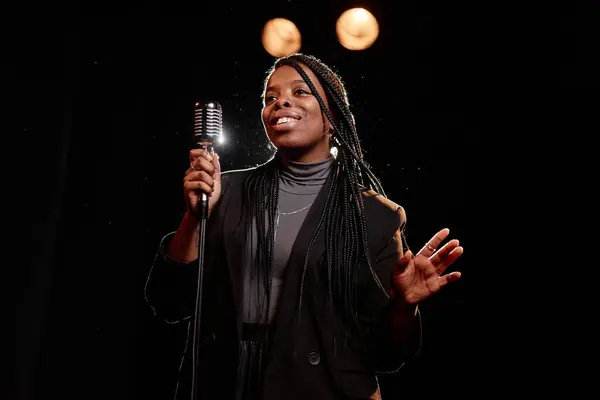Waist up portrait of elegant Black woman performing on stage with microphone against dark background copy space