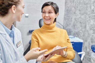 Diş kliniğinde dişçiyle konuşan ve elinde tablet olan gülümseyen olgun bir kadının portresi.