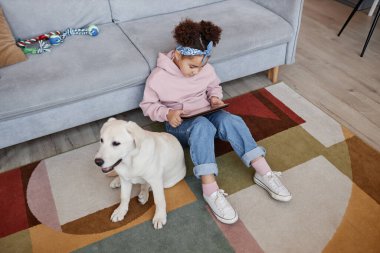 Tablet kullanan sevimli siyah bir kızın yüksek açılı portresi evde, şirin bir köpekle oturup, fotokopi çekiyor.