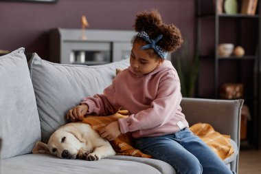 Evdeki kanepede uyuyan ve battaniyeyle kaplanmış şirin bir köpeğe bakan küçük siyah kızın portresi.