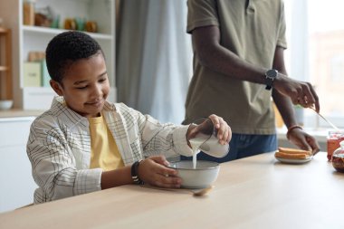 Kahvaltının fotokopi odasında mısır gevreği kasesine süt döken gülümseyen Afro-Amerikalı çocuğun portresi.