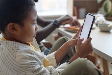Kahvaltı sırasında mutfak masasında otururken beyaz ekranlı akıllı telefon kullanan Afrikalı Amerikalı bir çocuğun yan görünümü.
