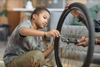 Babasıyla birlikte evde oturan siyah çocuğun bisiklet tekerleğini tamir edişinin yan görüntüsü.