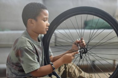Bisiklet tekerleklerini tamir eden, lastikleri kontrol eden Afrikalı Amerikalı bir çocuğun yan görünüm portresi.