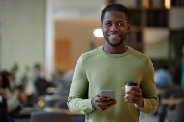 Modern siyah adamın portresi. Elinde akıllı telefon ve kahve fincanı var. Ofis binasının lobisinde kameraya gülümsüyor.