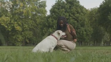 Güneşli bir günde şehir parkında yeşil çimlere çömelmiş, köpeğini okşayan ve ona ikramda bulunan bir zenci kız.