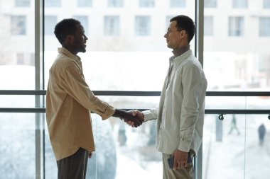 Siyahi ve Kafkas asıllı iki şık genç ofis binasının penceresinde el sıkışıyor.