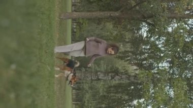 Melez köpeğiyle birlikte yeşil ağaçlar ve çimlerle parkta yürürken çekilmiş dikey bir beyaz kadın fotoğrafı.