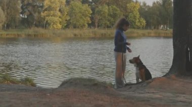 Genç bir kadın ve köpeği ormanın ortasında suyun yanında dikiliyorlar. Kadın hayvanına kıyak yapıyor.
