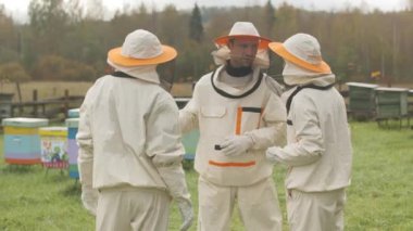 Arı kovanında koruyucu giysiler giymiş üç profesyonel arı yetiştiricisinin sohbet edip iş planlarını tartışırken orta boy bir fotoğraf.