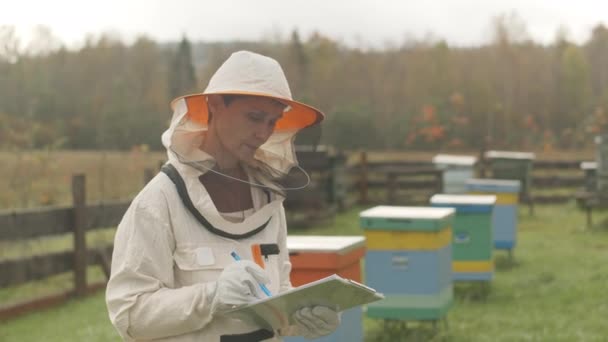 白种人女养蜂人头戴防护服 手持剪贴板 头戴相机 头戴中档慢幅肖像 工作天在农场花园 — 图库视频影像
