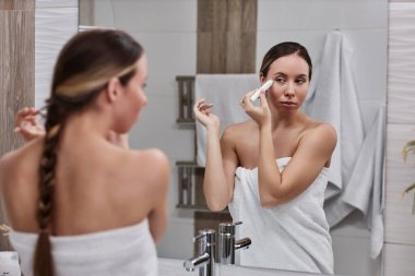 Banyoda göz kremi kullanan ve aynaya bakan genç bir kadının portresi.