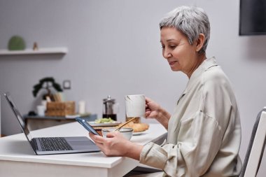 Modern son sınıf kadınının akıllı telefon kullanarak sabah kahvaltısının tadını çıkarırken çekilmiş yan görüntüsü.